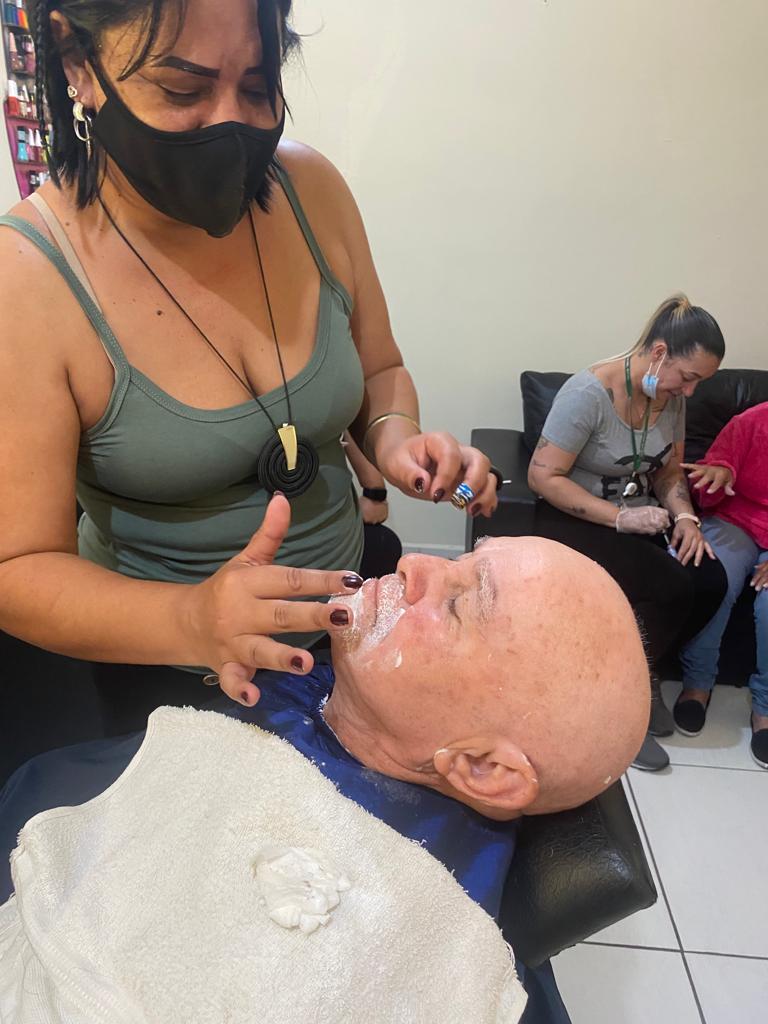 Na imagem, a cabeleleira faz a barba de um atendido do local, ela veste uma blusa verde e passa creme na barba do idoso. Ao fundo, uma mulher sentada em um sofá preto.